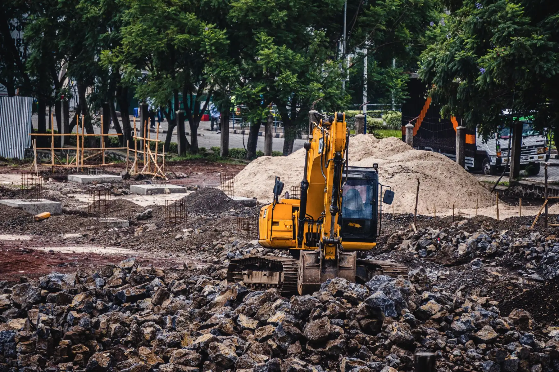 A yellow excavator working among piles of broken rock on a cleared site, with trees behind